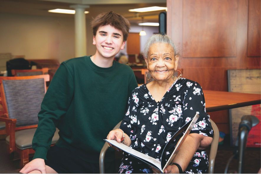 Young man in green sweater smiles beside an elderly woman in floral dress holding a book. They're in a warmly lit room with wooden decor.