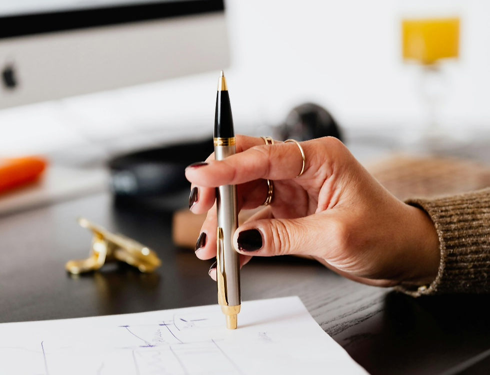 Hand with dark nails holds a pen over paper on a desk. Computer monitor and yellow mug in the blurred background. Cozy workspace vibe.