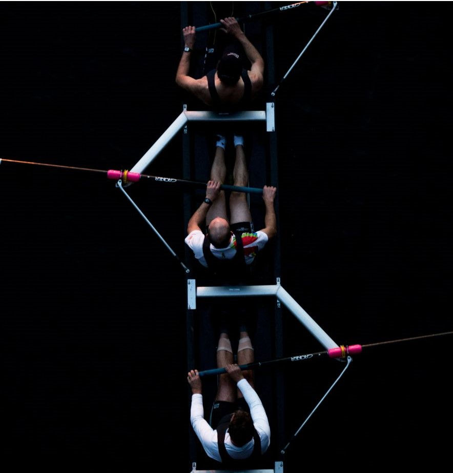 "Overhead view of two rowers in a rowing boat, with one wearing a sleeveless shirt and the other in a short-sleeved shirt, both holding oars in sync.