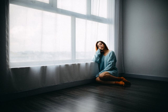Woman in a blue sweater and orange socks sits pensively by a large window in a minimal room, with soft light and muted tones.