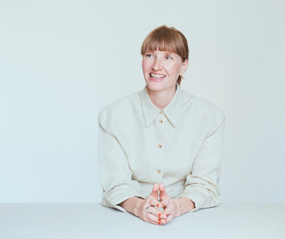 Woman with bangs smiling, sitting at a white table, wearing a light button-up shirt. Minimalistic background, bright and cheerful mood.