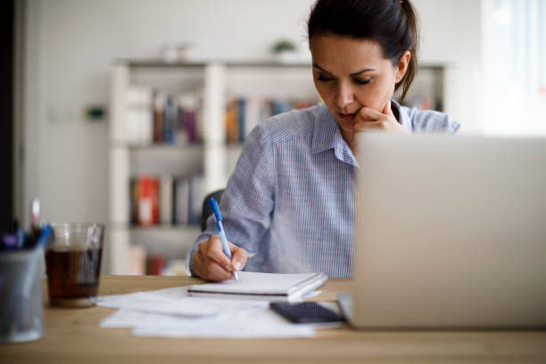 Woman writing notes at a desk with a laptop and documents. She's focused, in a casual setting with bookshelves in the background.