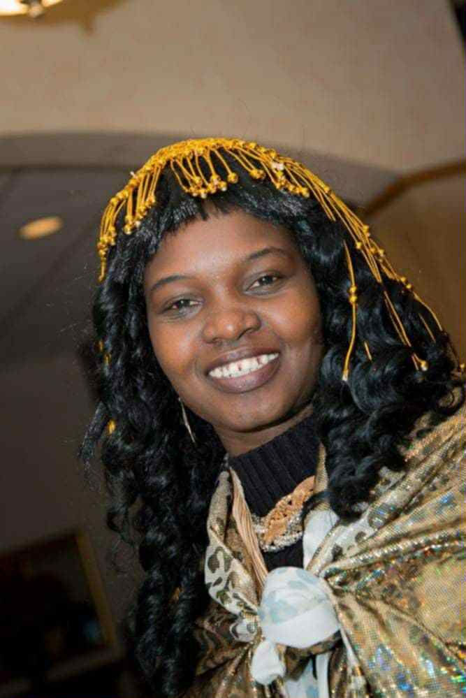 Smiling woman with curly hair adorned with gold beads, wearing a patterned shawl. Indoor setting with warm lighting.