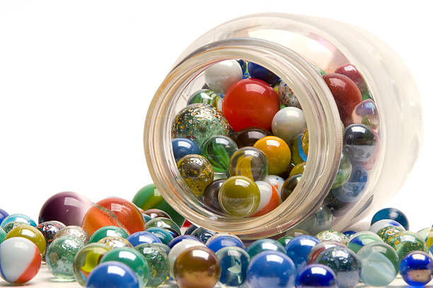 Colorful glass marbles spill from a clear jar onto a white surface, featuring vibrant reds, blues, and greens. Bright and playful mood.