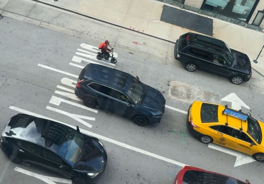 A person on a tiny motorbike rides in traffic alongside cars and a taxi on a city street.