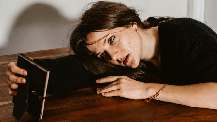 The image features a woman with a distressed expression, resting her head on a wooden table while holding a small black notebook.