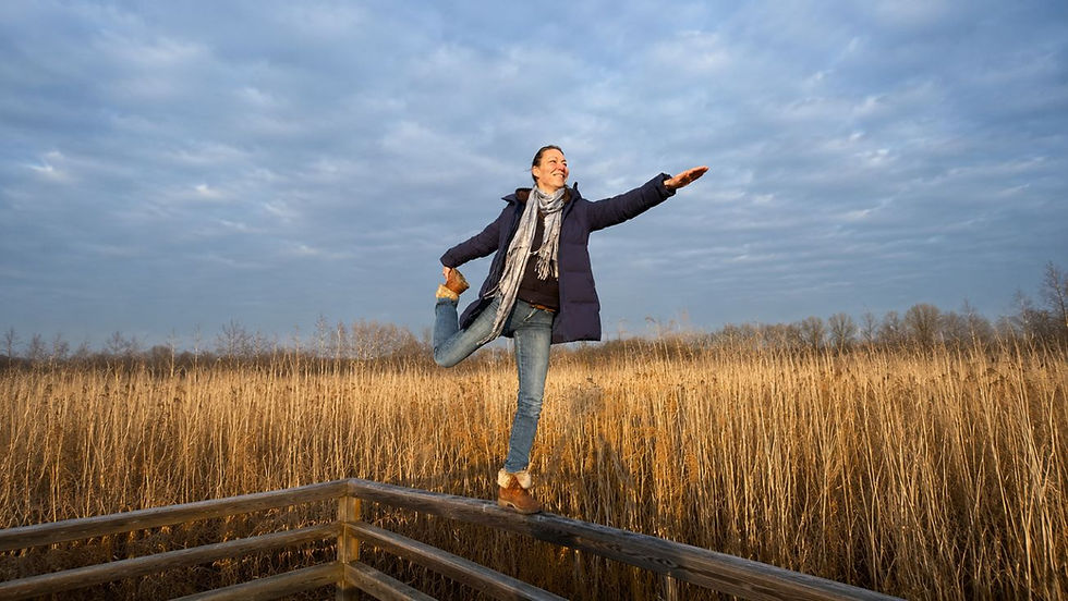 Woman joyfully poses on a wooden railing in a field of tall grasses under a cloudy blue sky, wearing a blue coat and scarf.