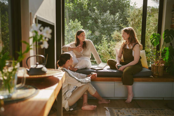 Three women chat on a sunny window seat surrounded by plants in a cozy room.