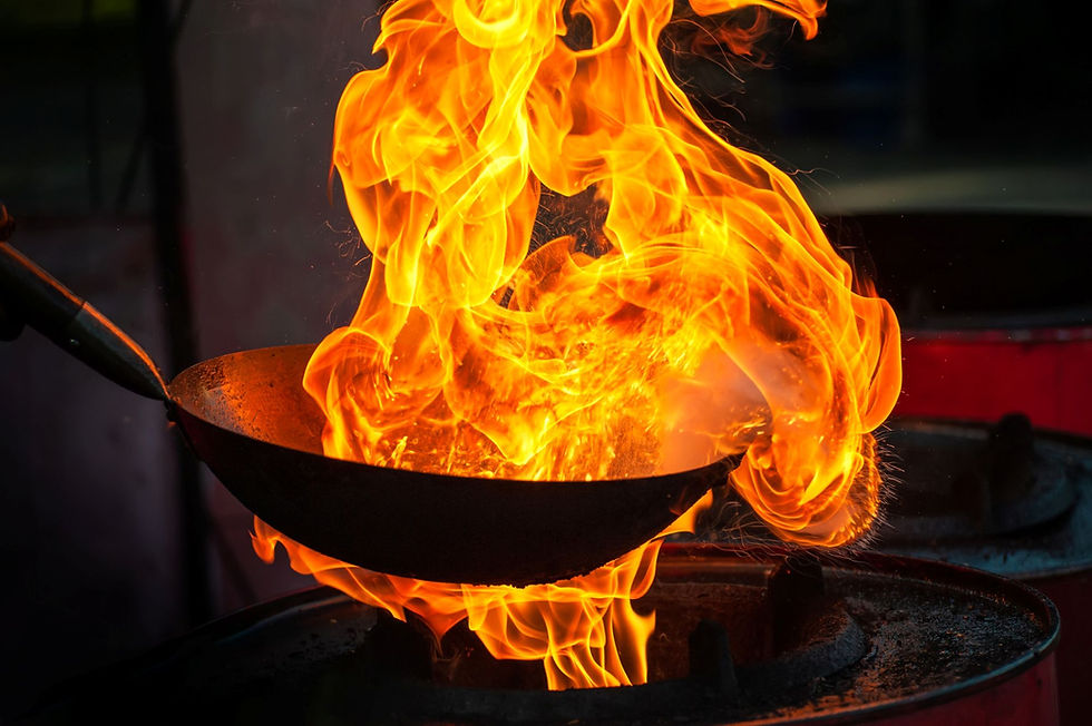 A wok on a stovetop erupts in bright orange flames, creating a dramatic, intense kitchen scene.