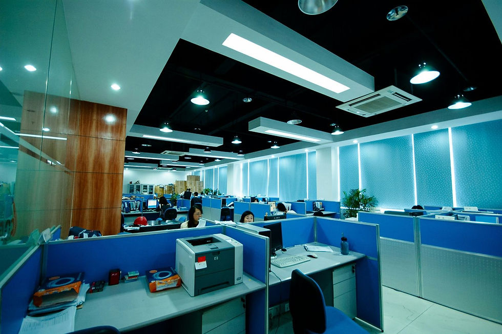 Modern office with blue cubicles, people working at desks, and overhead lights. A printer is on a desk in the foreground. Bright and tidy.