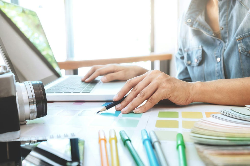 Hands using a laptop beside color swatches and pencils in a bright room. A camera and pens add creative elements to the scene.