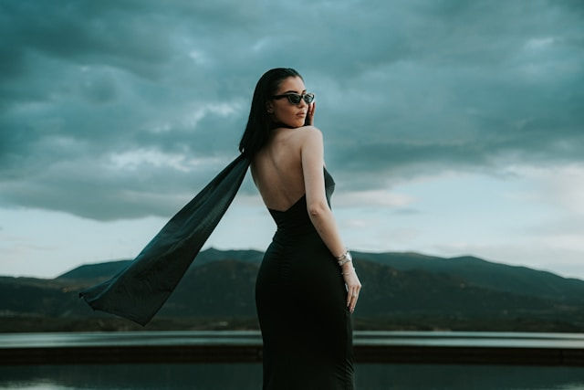 A woman in a black dress and sunglasses stands with a long black scarf flowing behind her, against a backdrop of mountains and a stormy sky.