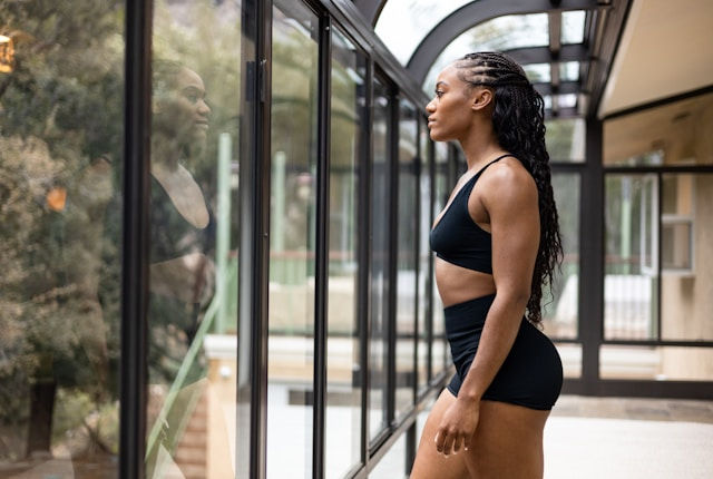 A woman in athletic wear stands confidently in front of a glass wall, gazing at her reflection with determination.