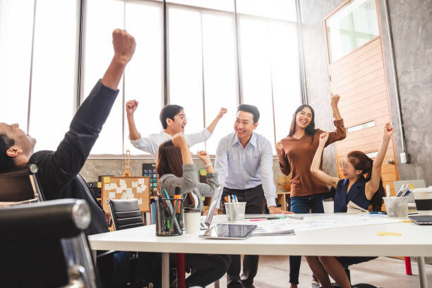 Office team celebrates success around a table, smiling, arms raised. Bright room, whiteboard in background. Energetic, joyful atmosphere.