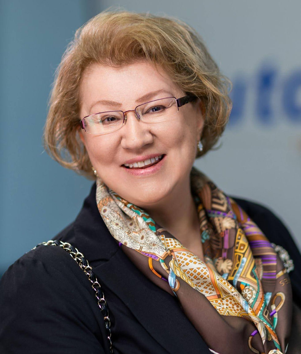 A smiling person with short, light hair wears glasses and a patterned scarf. The background is blurred, with blue and cream tones.