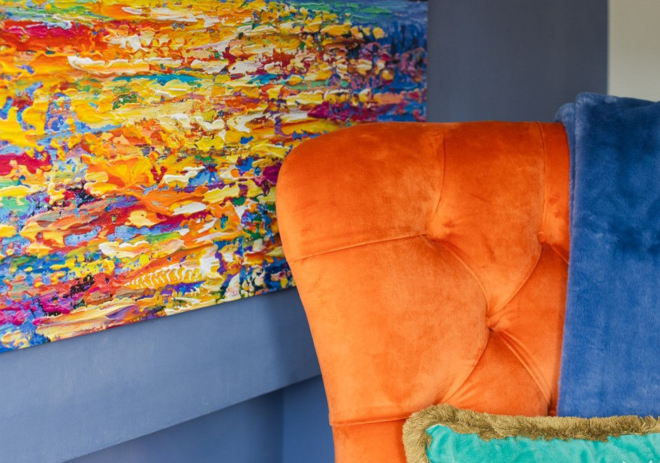 Colorful abstract wall art behind an orange tufted chair with a blue throw, in a bright modern room.