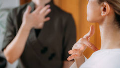 Two people in therapeutic gestures; one in focus, touching neck, wearing white. Background person in brown, soft light, calm setting.