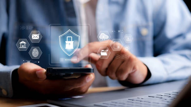 Person in blue shirt uses a smartphone beside a laptop. Digital icons, including a lock, hover above their hand, suggesting cybersecurity.