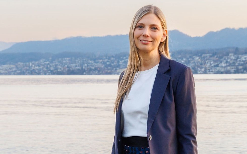 Smiling woman in a blue blazer stands by a calm lake, with mountains and a town in the background, under a soft, pastel sky.
