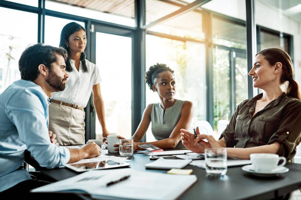 The photo shows a group of four professionals engaged in a meeting, with two women standing and two men sitting at the table, actively discussing and exchanging ideas.