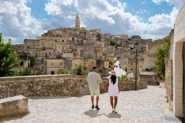 Couple holding hands walks on a sunlit stone pathway in an ancient town. Cloudy blue sky and historic buildings create a serene setting.