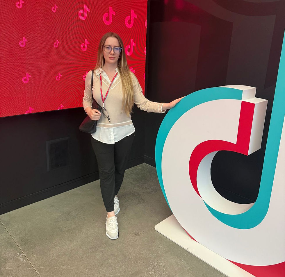 Woman in glasses stands next to large TikTok logo. Background features red pattern with TikTok symbols. Casual attire, neutral expression.