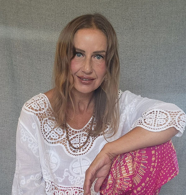 Woman in white lace top and pink patterned pants sits against a gray wall, smiling softly at the camera.