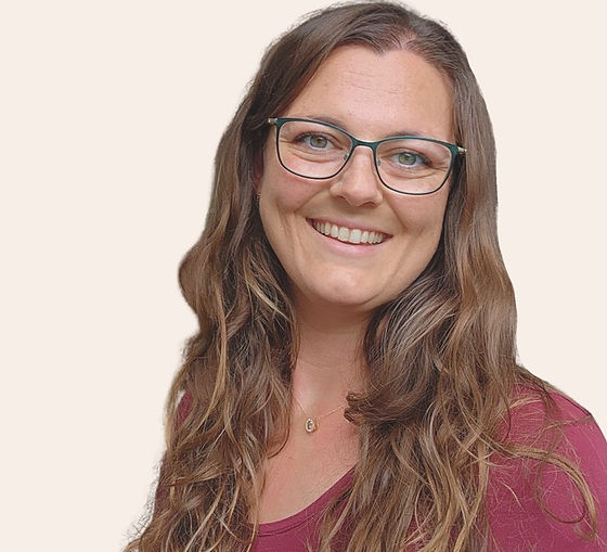 Smiling woman with glasses and long wavy brown hair, wearing a magenta top against a plain light background.