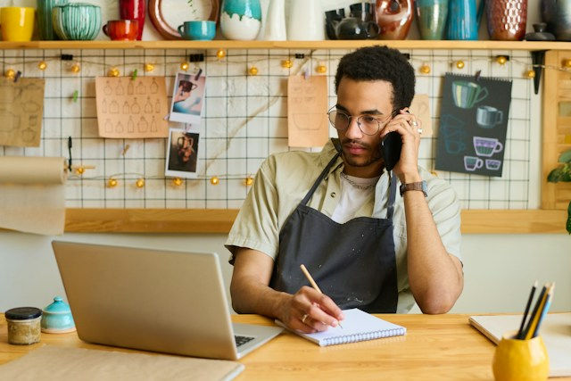 Young man in apron talks on phone while writing at a laptop in a cozy café workshop with hanging mugs and warm lights.