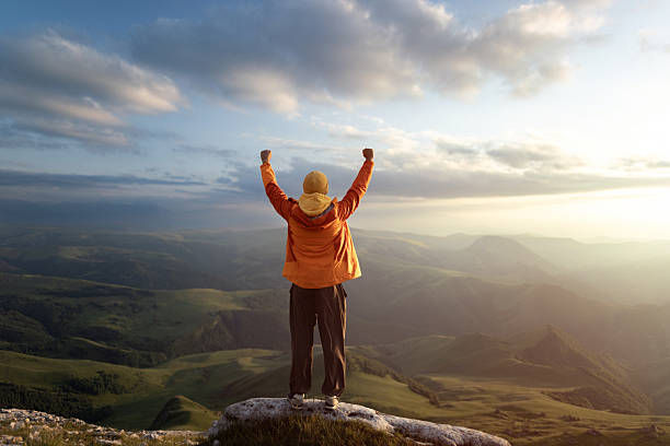 Hiker in orange jacket stands on a cliff with arms raised at sunrise over misty mountain valleys