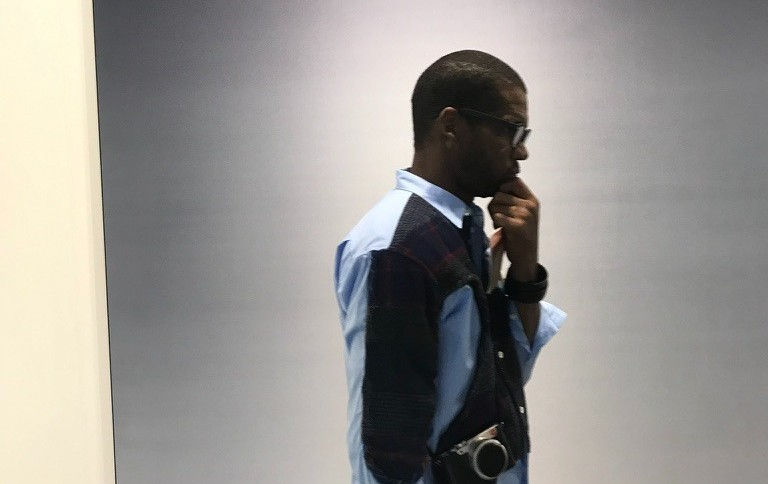 Profile of a thoughtful man in sunglasses and a blue shirt, hand on chin, standing against a plain gray wall with a camera at his side