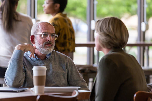 Two people engage in conversation at a cafe table, one holding a coffee cup. Others are visible in the blurred background, creating a casual mood.