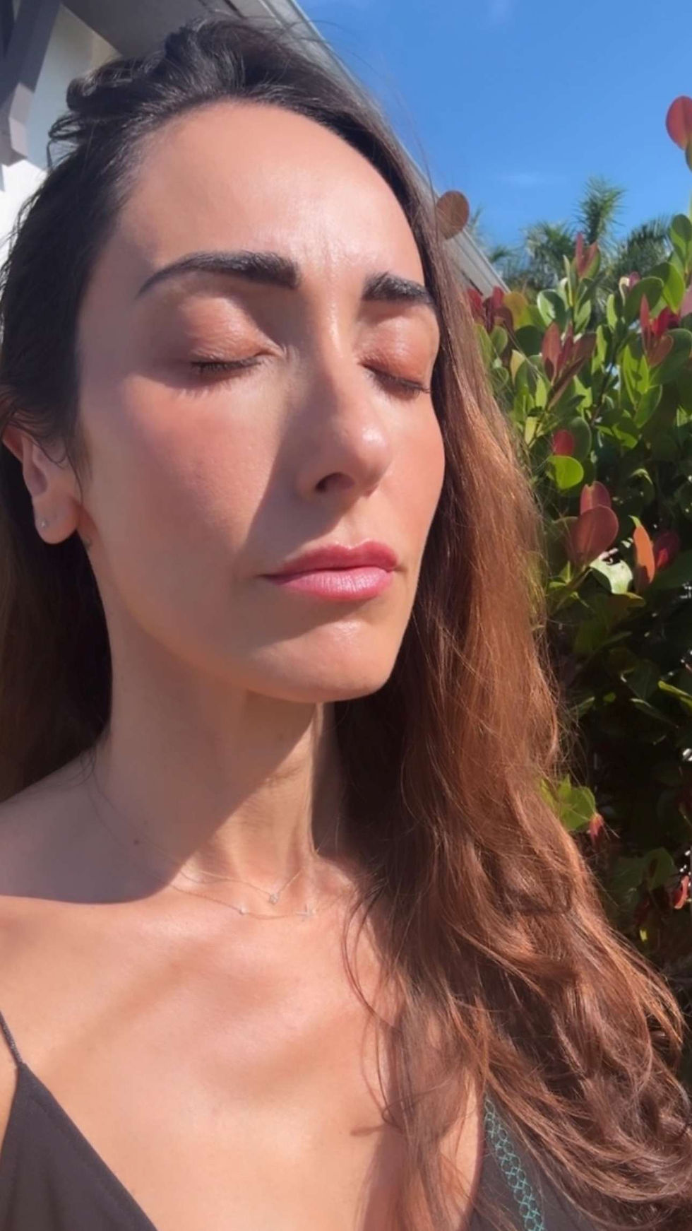 Woman with closed eyes basks in sunlight, surrounded by lush green and red leaves. Clear blue sky in the background. Calm expression.