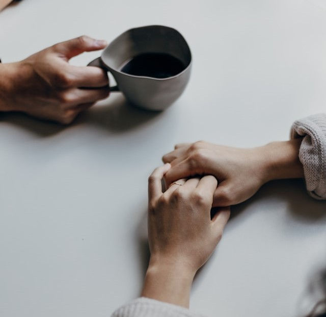Hands hold each other affectionately on a light table. One person holds a gray cup with dark liquid, creating a calm and intimate mood.