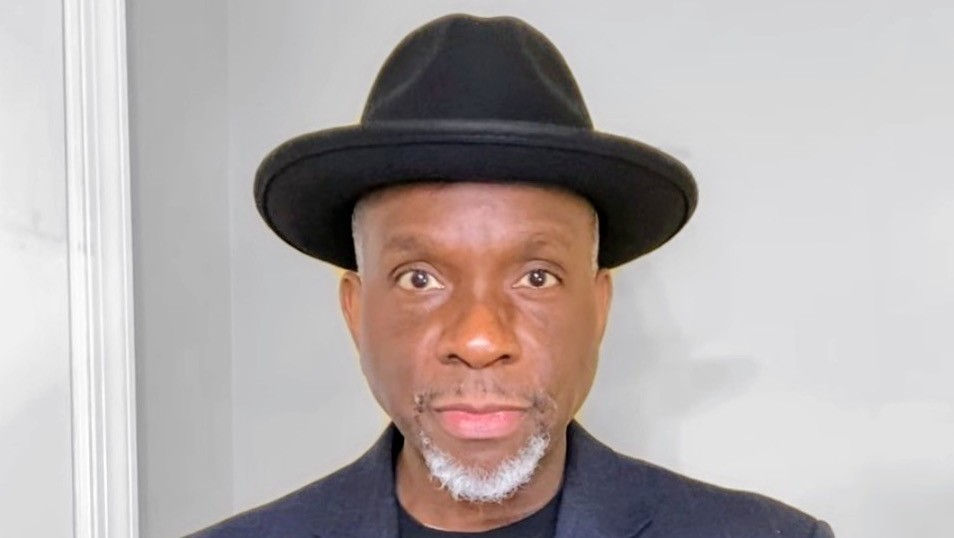 Portrait of a man in a black hat and dark jacket against a light gray wall, looking straight at the camera with a neutral expression.