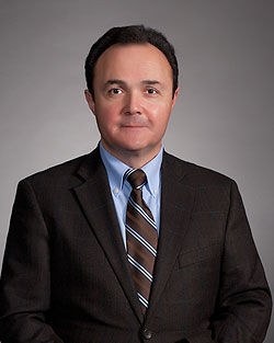 Man in a dark suit with a striped tie, blue shirt, neutral expression, gray backdrop, formal setting. No visible text.