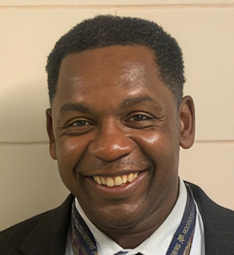 Smiling man in a suit with a patterned tie, against a light-colored wall. Happy expression, indoor setting.