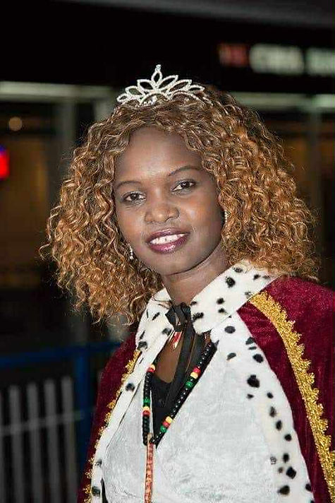 Woman wearing a crown and red velvet cape smiles in a well-lit setting. Her curly hair and pearl tiara stand out against the blurred background.