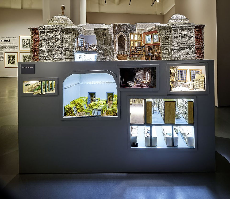 Museum exhibit of a detailed building model with lit cutaway rooms and photos on a gray display in a gallery.