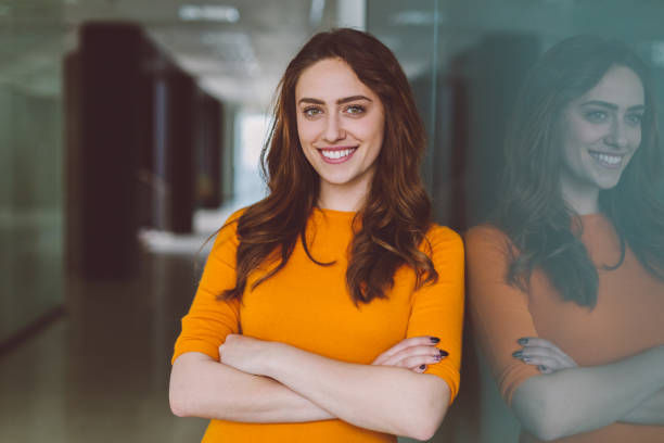 Smiling woman in an orange sweater with arms crossed, standing indoors against a glass wall. Office setting, bright and confident mood.