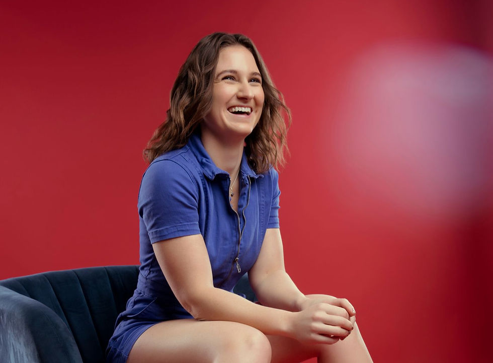 Smiling woman in a blue dress sits on a dark chair against a vivid red backdrop, laughing warmly.