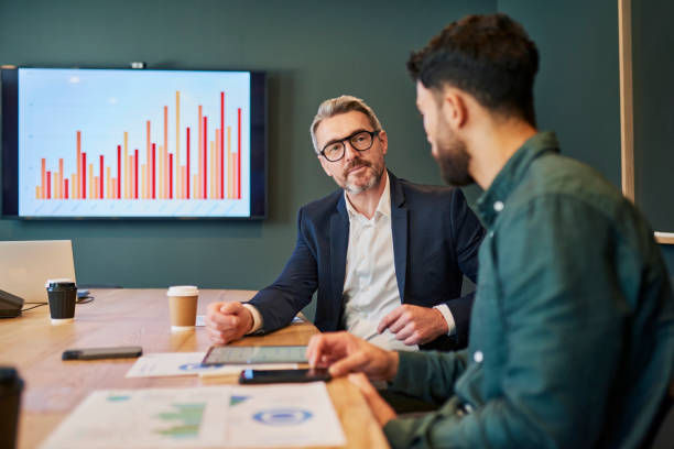 Two men in a meeting room discuss graphs on a screen. Documents and coffee cups sit on the table. The mood is professional.