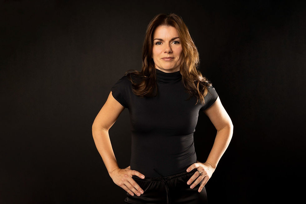 Confident woman in a black top stands with hands on hips against a dark studio background.