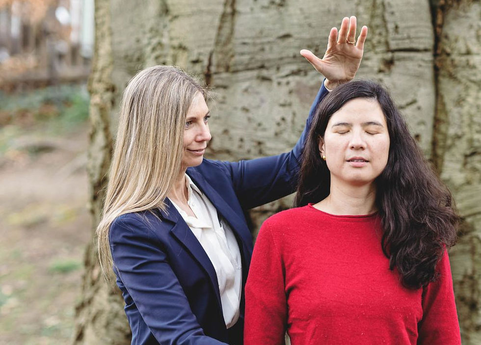 A woman in a dark blue blazer places her hand above the head of another woman wearing a red sweater. The woman in red has her eyes closed. They appear to be outdoors, near a tree trunk.