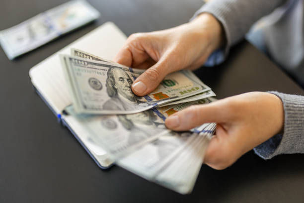 Hands counting 100-dollar bills over an open notebook on a dark surface; focus on the money, creating a mood of financial management.