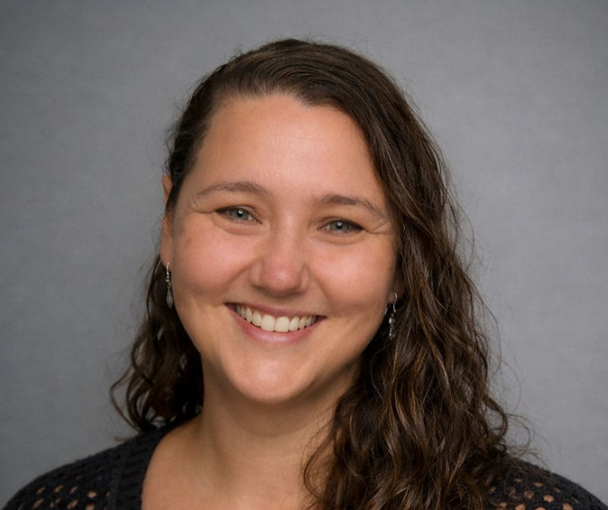 Smiling person with long, curly hair, wearing a dark top. Gray background. Mood is joyful and welcoming.