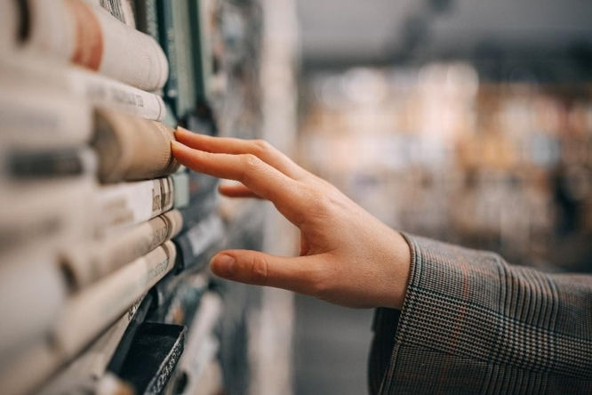 Hand browsing book spines on a shelf in a bookstore or library, with a soft blurred background and a calm, curious mood.