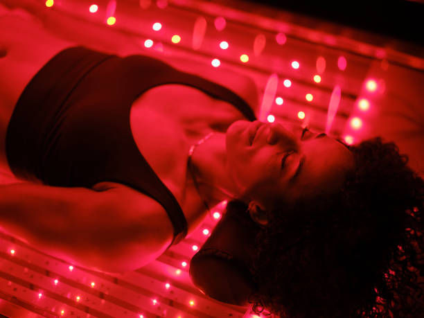 A person lying on a red LED light therapy bed, eyes closed, looking relaxed. The scene is bathed in vibrant red light.