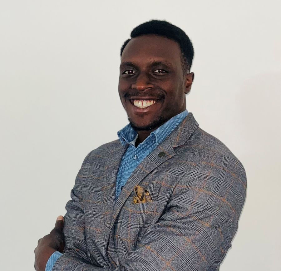Smiling man in a gray plaid blazer and blue shirt stands against a plain white background.