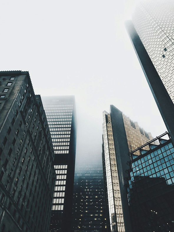 Tall skyscrapers shrouded in fog reach into a cloudy sky. Reflective glass and concrete create a moody, urban atmosphere.